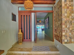 Un pasillo con una puerta de madera, un jarrón tejido y una alfombra estampada que conduce a una habitación con vistas a la playa.