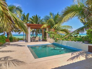 Zona de piscina con un pérgola, palmeras y vista al océano.