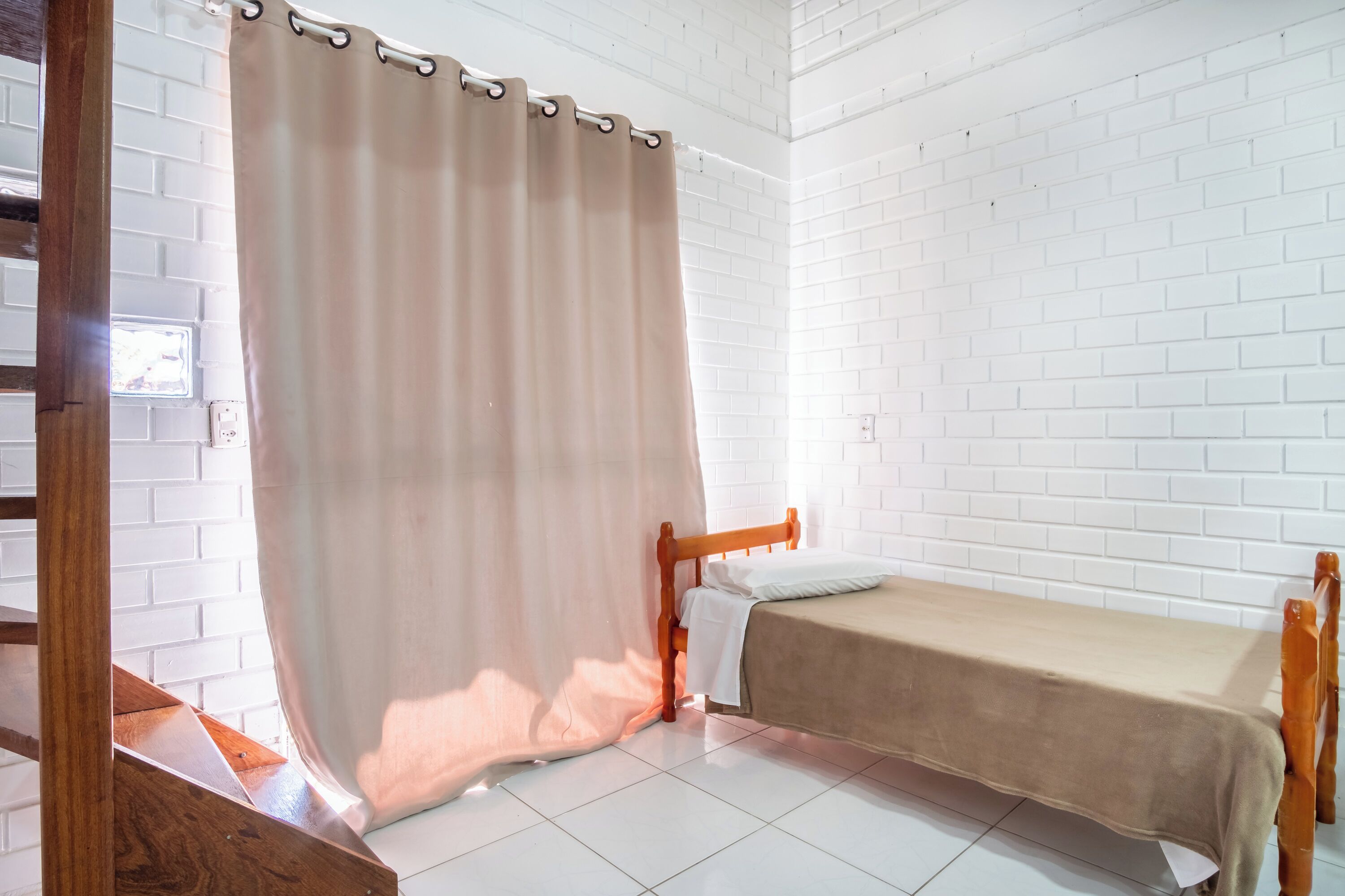 A small, clean room with white brick walls, a wooden bed frame, and a single bed with a beige cover.