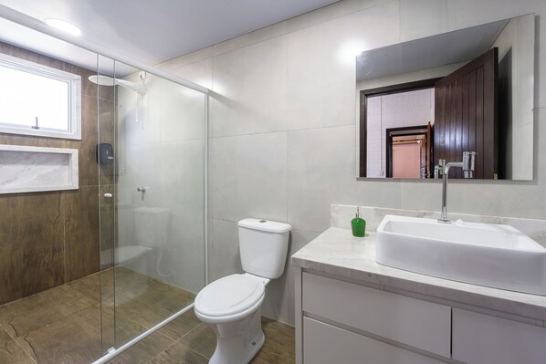 Un baño moderno con ducha envuelta en vidrio, inodoro blanco, lavamanos de mármol y tocador de madera.