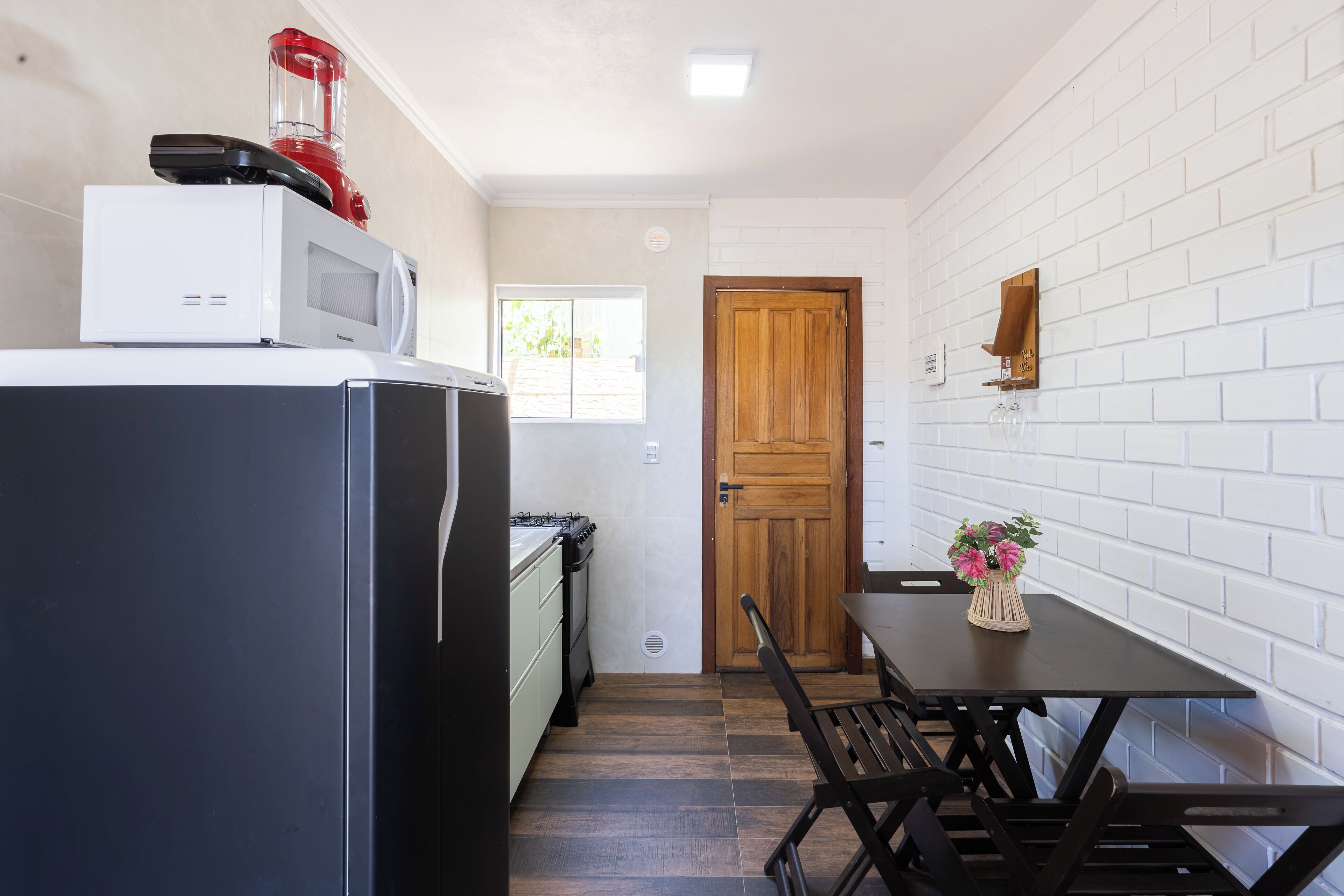 Cocina compacta con refrigerador, microondas y estufa. Se observa una puerta de madera y una pared de ladrillo blanco. Hay una mesa de comedor con sillas.