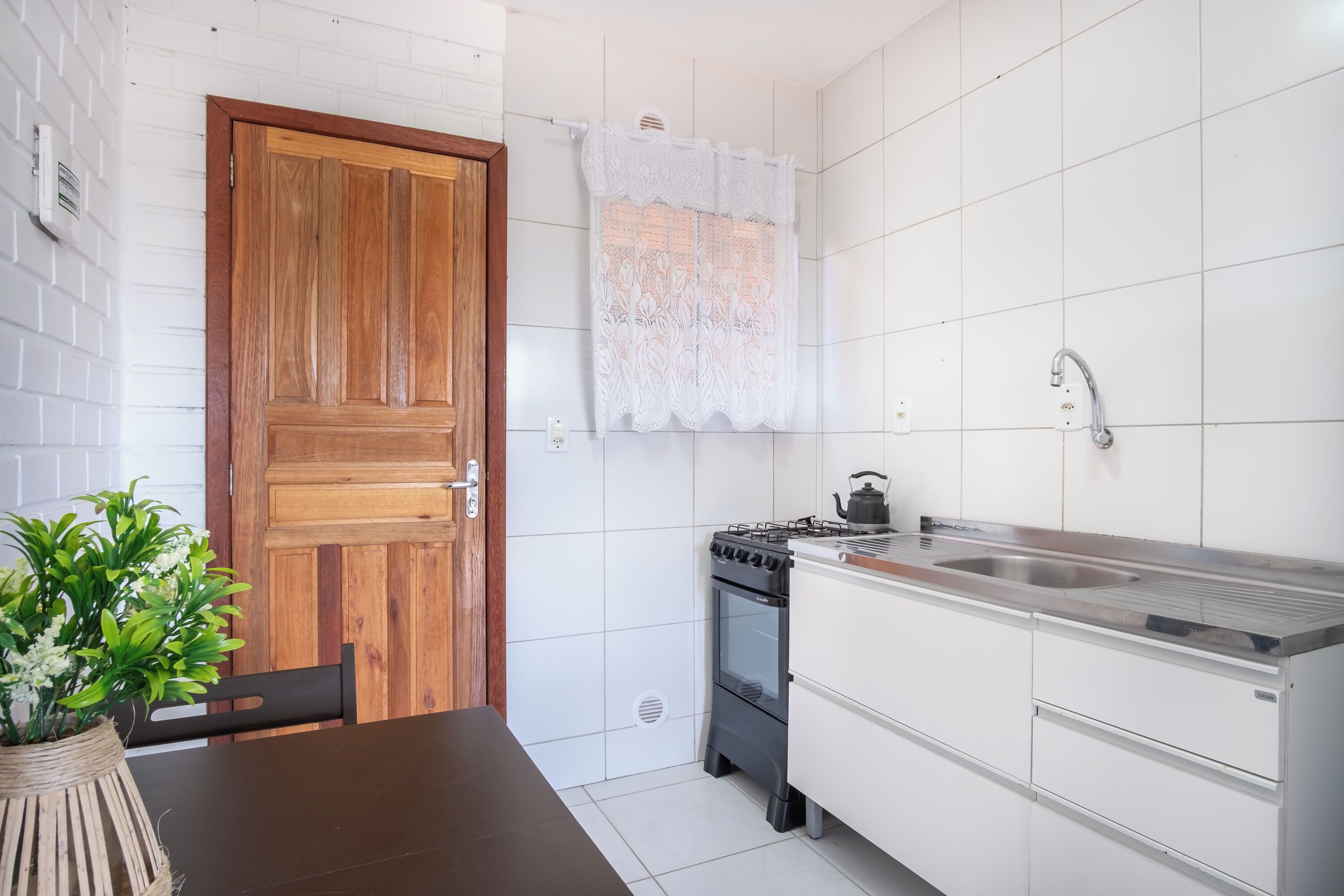 Una cucina con pareti piastrellate di bianco, una porta in legno, un fornello, un lavandino e un vaso con piante verdi.