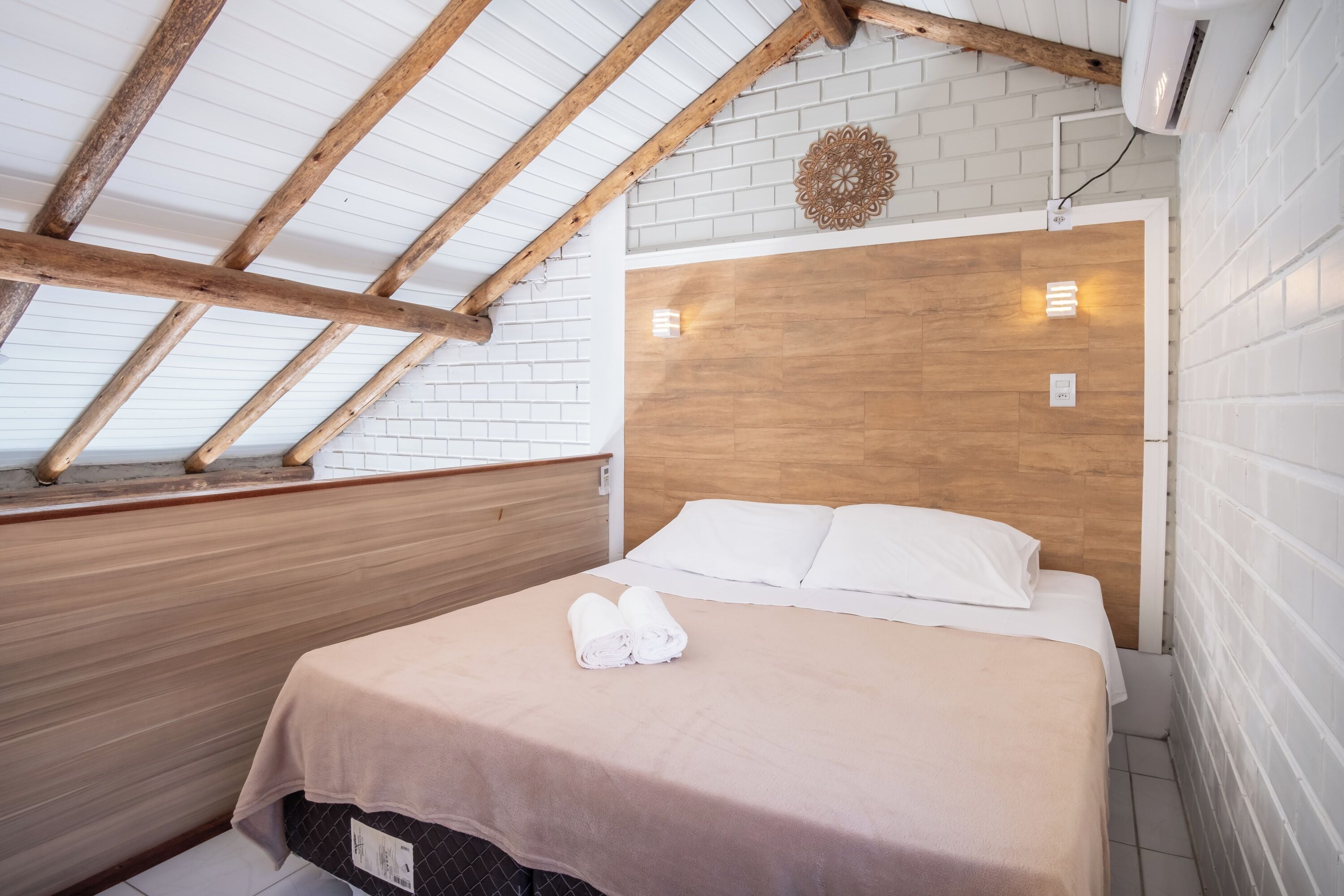 A bedroom with a sloped ceiling, wooden beams, a bed with white linens, and a wooden headboard.