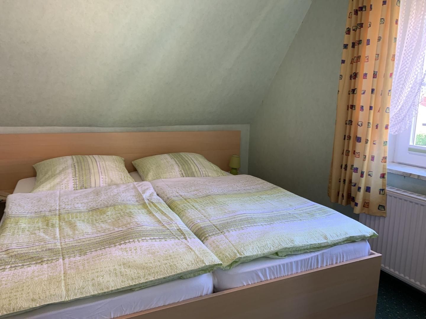 Een enkel bed met een houten hoofdbord, een bedlamp en een raam met gordijnen.