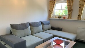 A modern living room with a grey sectional sofa, a glass-top coffee table, and a window with curtains. - Wiesenblick - Idyllic and quiet location, family-friendly (Grube)