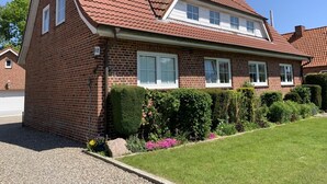 Et tosset hus i mursten med røde tegltag, hvide vinduer og en velholdt forhave med hække og blomster.
