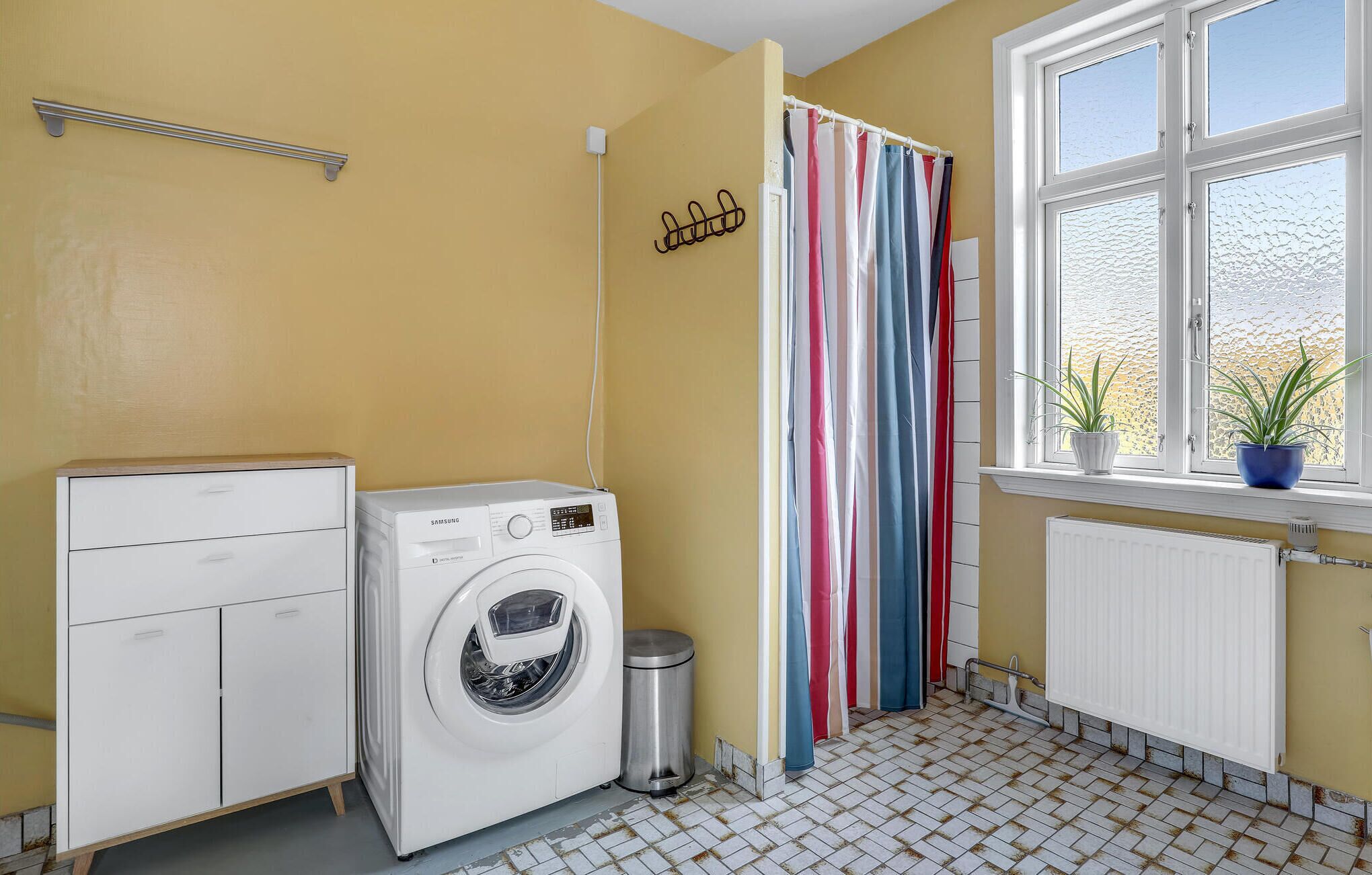 Una lavandería con lavadora, tendedero, radiador y ventana con planta.