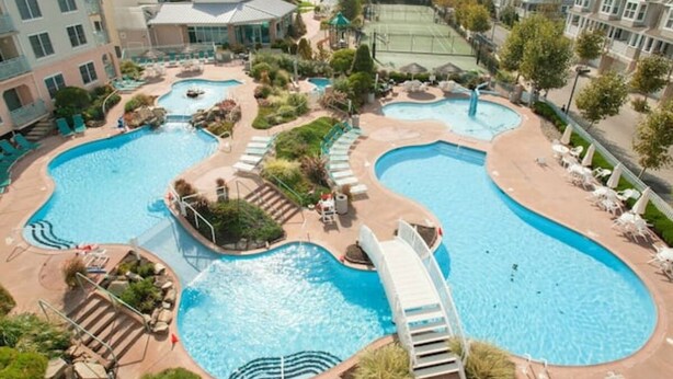 Un resort con varias piscinas, cancha de tenis y una variedad de sillas y sombrillas de descanso.