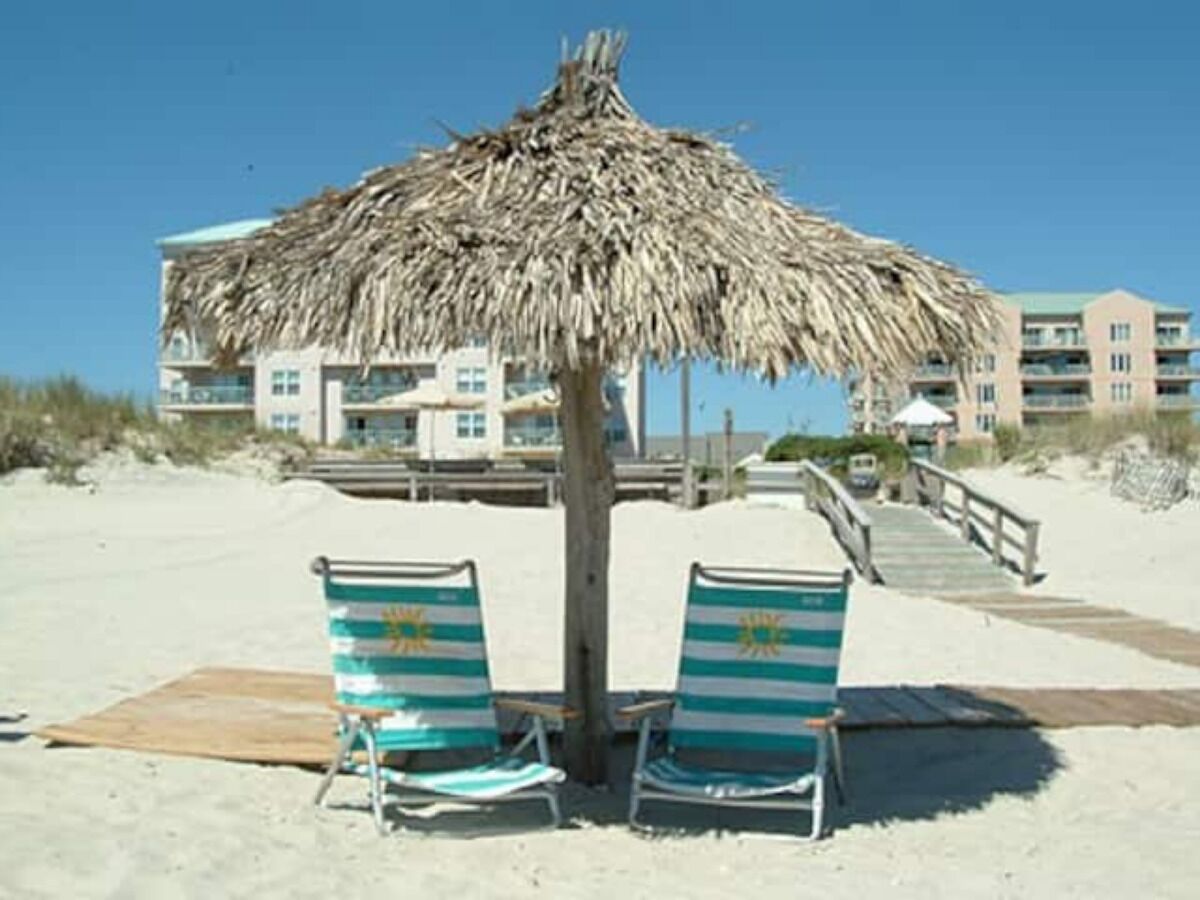 A beach with two striped beach chairs under a thatched umbrella, a wooden deck, and a building in the background.
