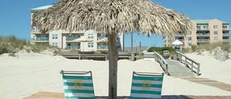 A beach with two striped beach chairs under a thatched umbrella, a wooden deck, and a building in the background.