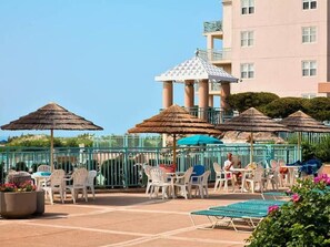 Área de varanda externa com mesas, cadeiras, guarda-sóis e piscina.