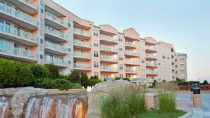 Un hotel de varios pisos con balcones, un jardín ajardinado con una cascada pequeña y un sendero pavimentado.