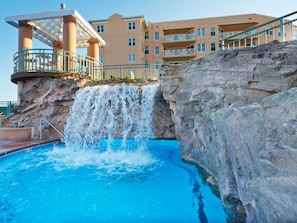 Hotel com piscina, apresentando uma cascata, área coberta com corrimão e um edifício de vários andares ao fundo.