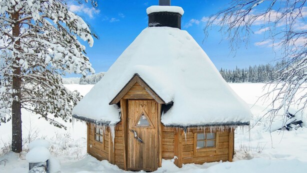 A wooden cabin with a snow-covered roof and chimney, surrounded by a snowy landscape.