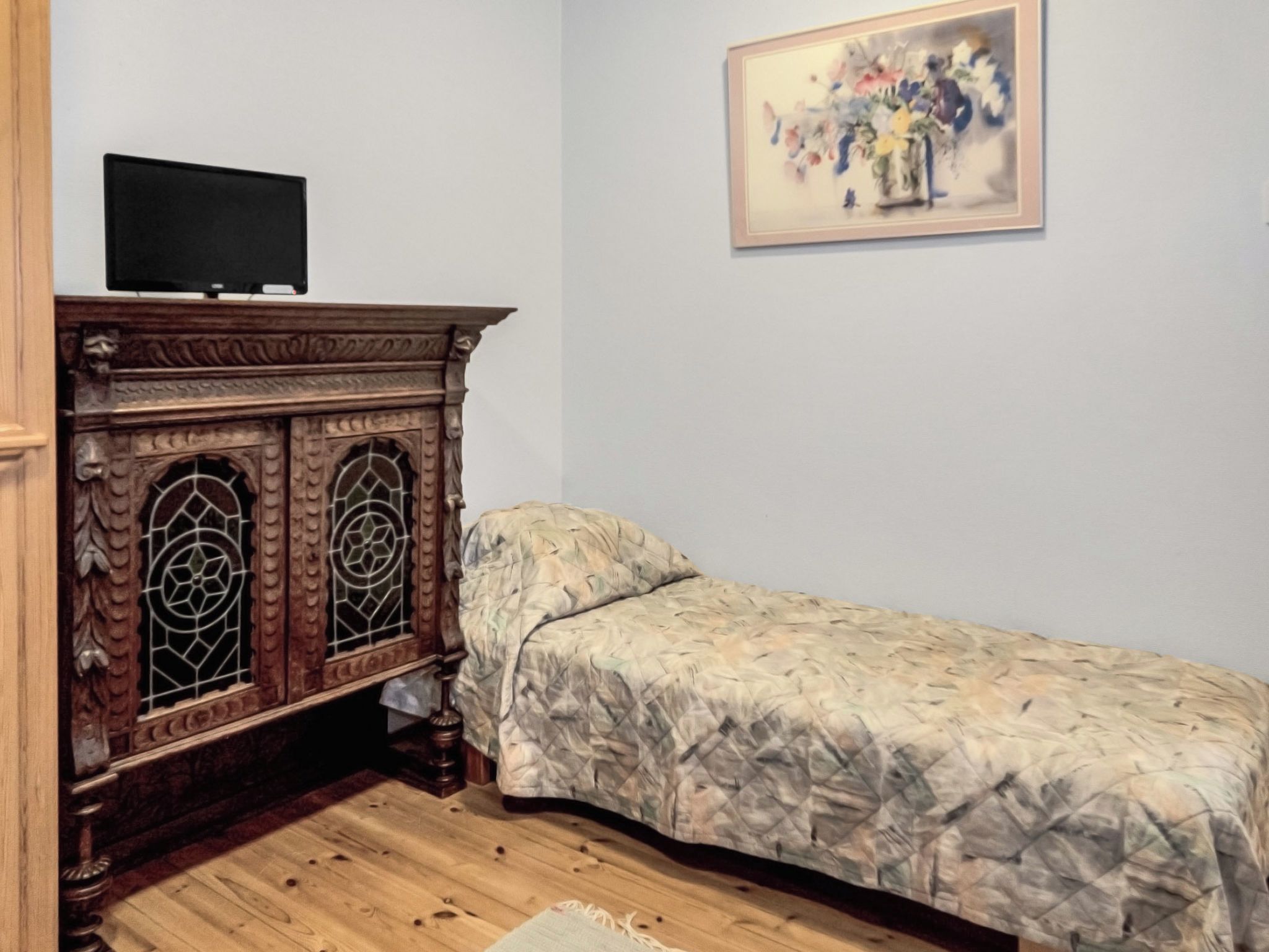 Een slaapkamer met een houten kast, een bed met een geborduurde spreideken, een televisie op de kast, en een schilderij aan de muur.
