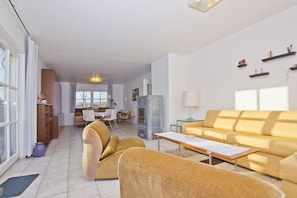 Ein geräumiges Wohnzimmer mit einem gelben Sofa, einem Couchtisch und einem Kamin. Im Hintergrund befindet sich ein Essbereich mit Tisch und Stühlen.