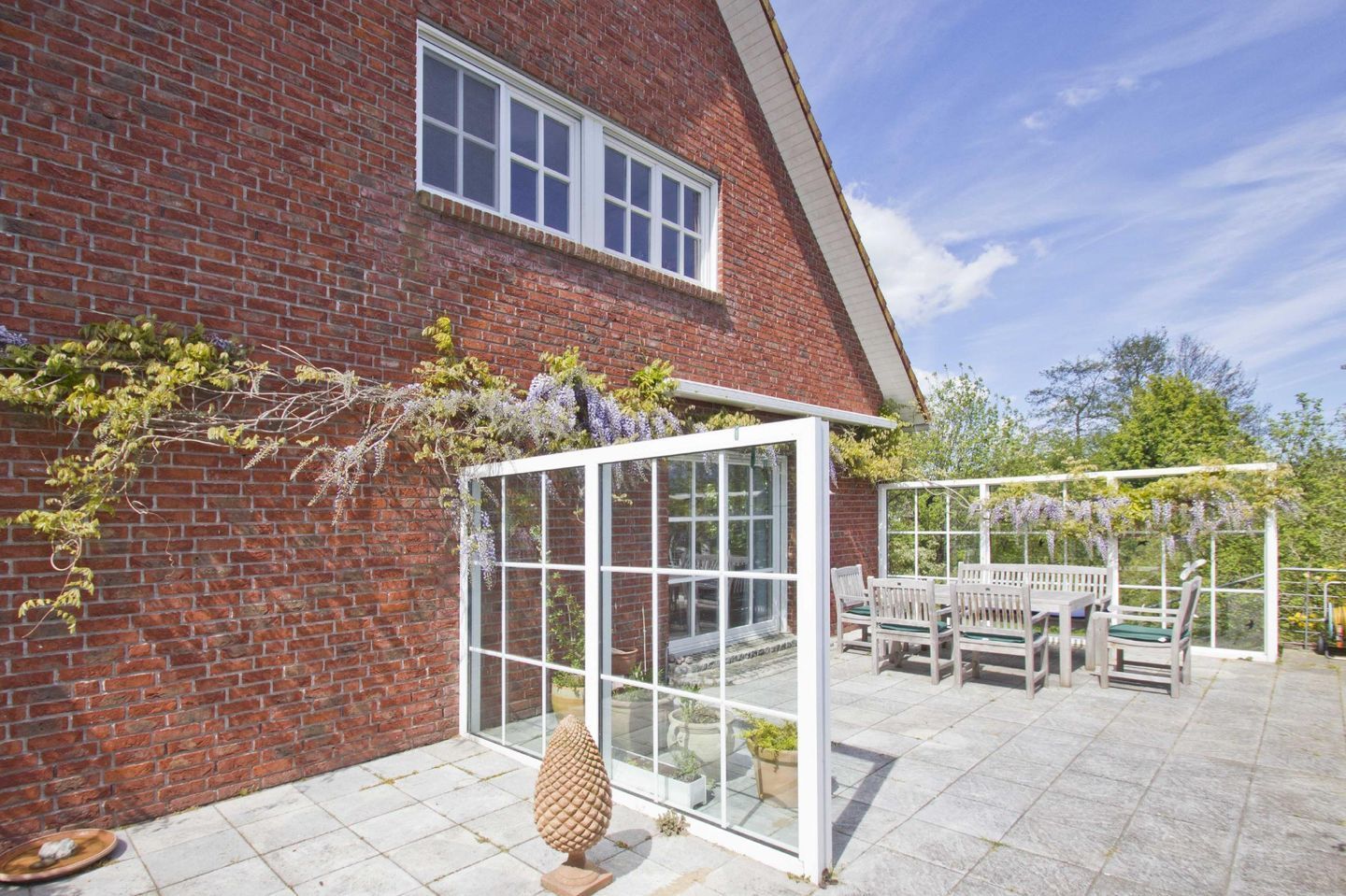 Une maison en briques avec une véranda en verre, une terrasse avec une table et des chaises, et une sculpture décorative en pierre.
