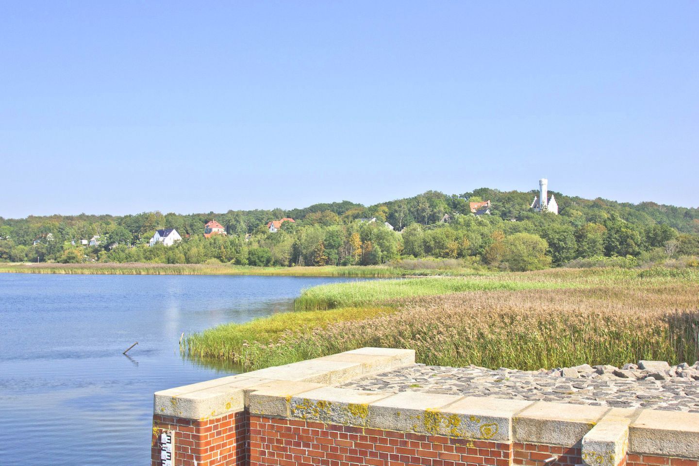A lakeside view with a brick wall in the foreground, a calm lake, and houses on a hill in the background.