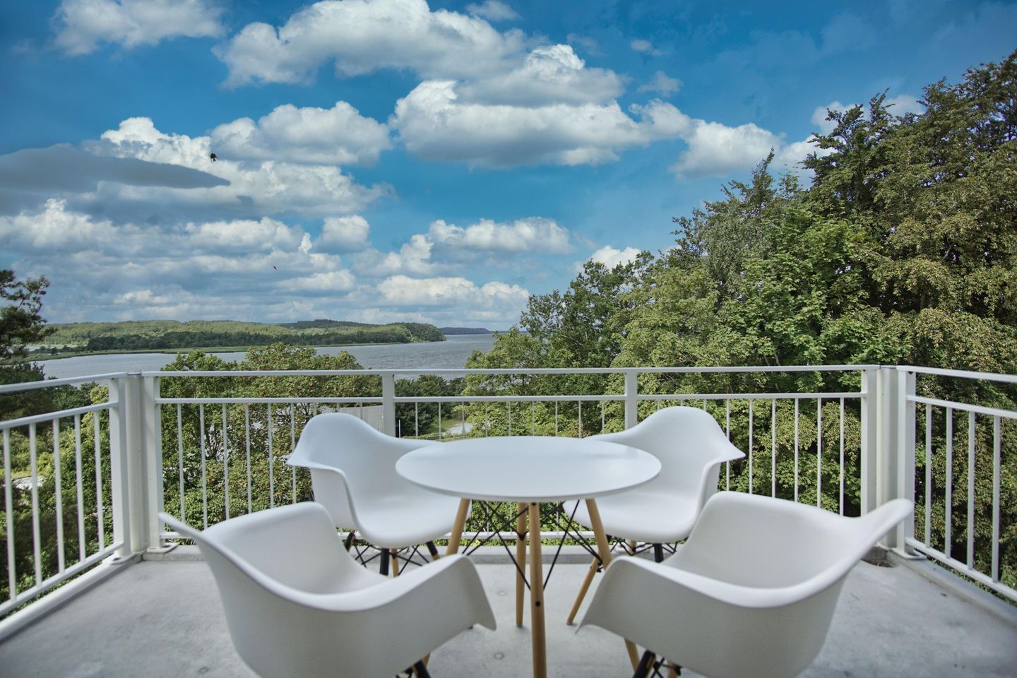 A balcony with white chairs and a table, overlooking a lake and forested hills.
