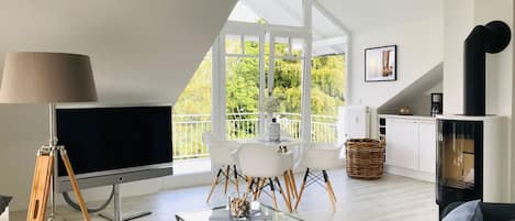 A modern living room with a large TV, a sofa, a dining table with chairs, and a kitchen area with a stove.