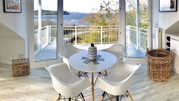 A modern room with a round table and four white chairs, large windows with a view of the sea, and a wicker basket on the floor. - Modern vacation apartment with balcony, sea view - Ferienresidenz Zwei BoddenFeWo 1-5 (Lietzow)