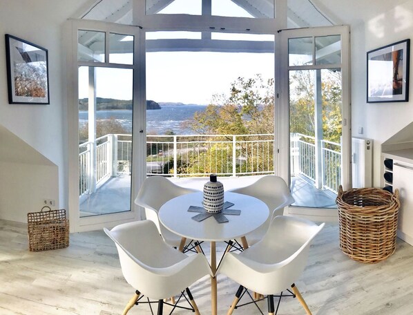 A modern room with a round table and four white chairs, large windows with a view of the sea, and a wicker basket on the floor.