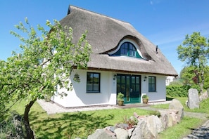 Ein traditionelles Haus mit einem Strohdach, einer grünen Tür, weißen Wänden und einem kleinen Garten mit Baum und Steinen.
