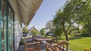 Un patio al aire libre con una mesa y sillas de madera, una estructura de techo de paja y una vista de una casa blanca con techo de paja al fondo.