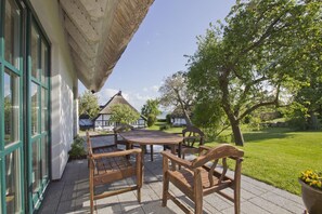 An outdoor patio with a wooden table and chairs, a thatched roof structure, and a view of a white house with a thatched roof in the background. - Gutshof Ostseeblick - beachfront idyll by the sea, private bathing area, beach chair (Putbus)