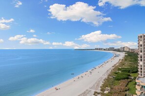 A coastal beach with a long stretch of sand, a clear blue sea, and a few buildings along the shore.