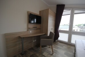 A hotel room with a wooden desk, a TV mounted above it, a chair, and a large window with a view of buildings and water. - With a view of the sea and a balcony, access to the pool and sauna - Sea View Apartment 66 - (Göhren)