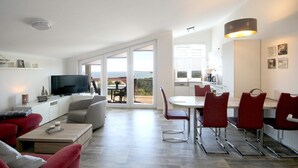 Um apartamento moderno com sala de estar, área de jantar e cozinha. Há uma televisão grande, uma mesa de jantar com cadeiras e uma cozinha com vista.
