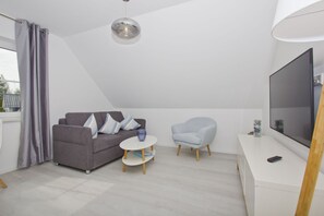 Ein modernes Wohnzimmer mit grauem Sofa, einem hellblauen Sessel, einem kleinen weißen Tisch und einem großen Flachbildfernseher.