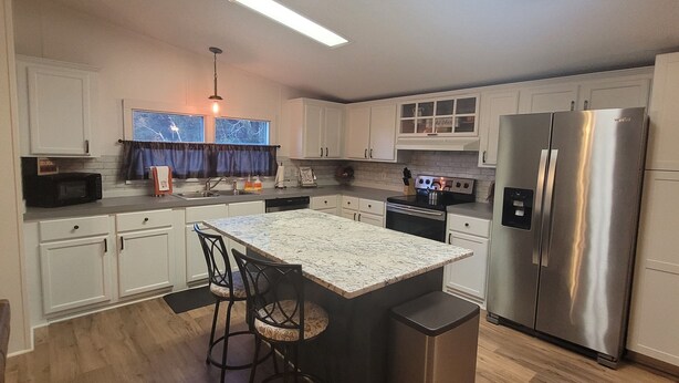 A modern kitchen with white cabinets, a stainless steel refrigerator, a built-in oven, and a granite countertop island with bar stools. - The DW - Spacious and Cozy (Columbia)