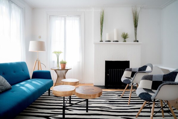 Una sala moderna con un sofá azul, un sillón estampado, una mesa de centro redonda de madera, una lámpara de pie y una chimenea con adornos.