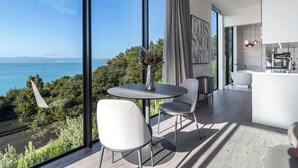 A modern interior with a dining area, a kitchen, and a large window overlooking a scenic view. - Omana (Waiheke Island)