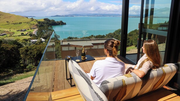 A couple seated on a balcony overlooking a scenic view of a lake and distant mountains. - Omana (Waiheke Island)