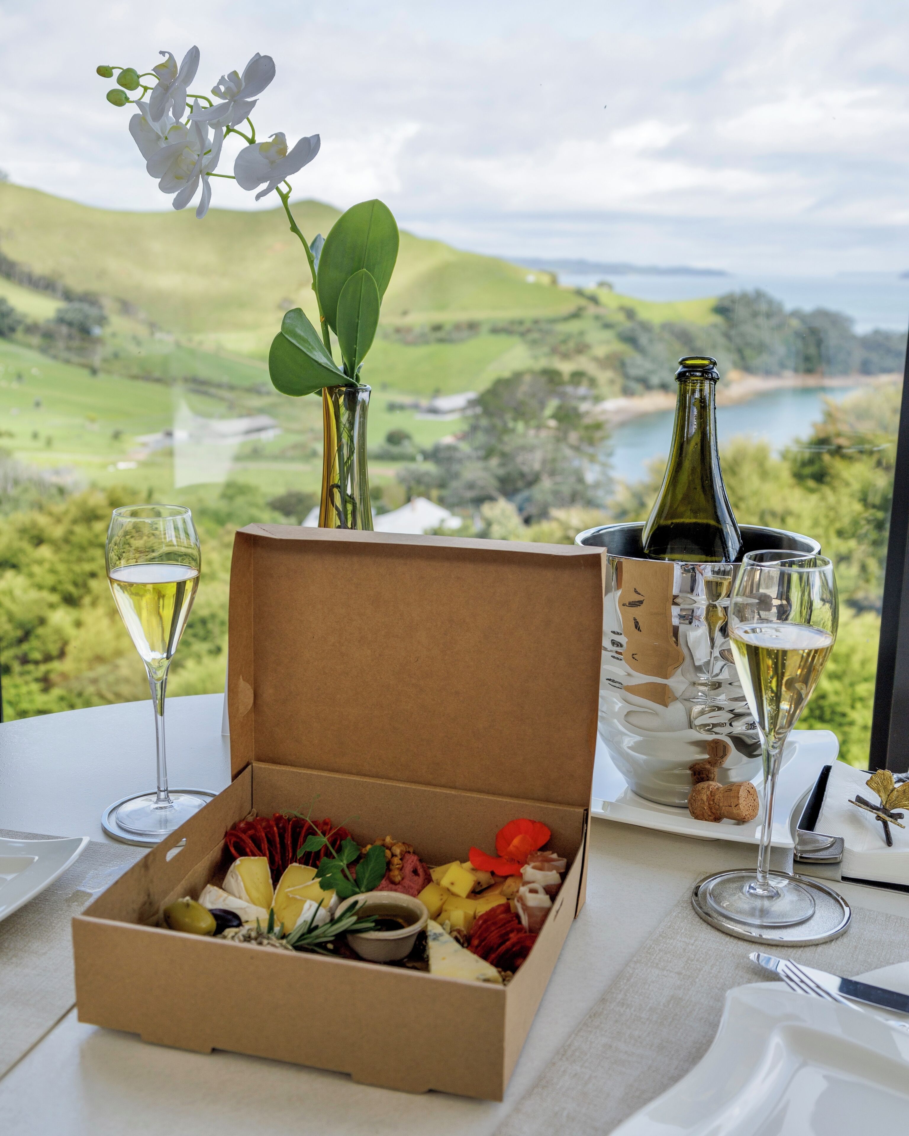 A dining table with a box of assorted cheeses, a bottle of wine, and two glasses of white wine.