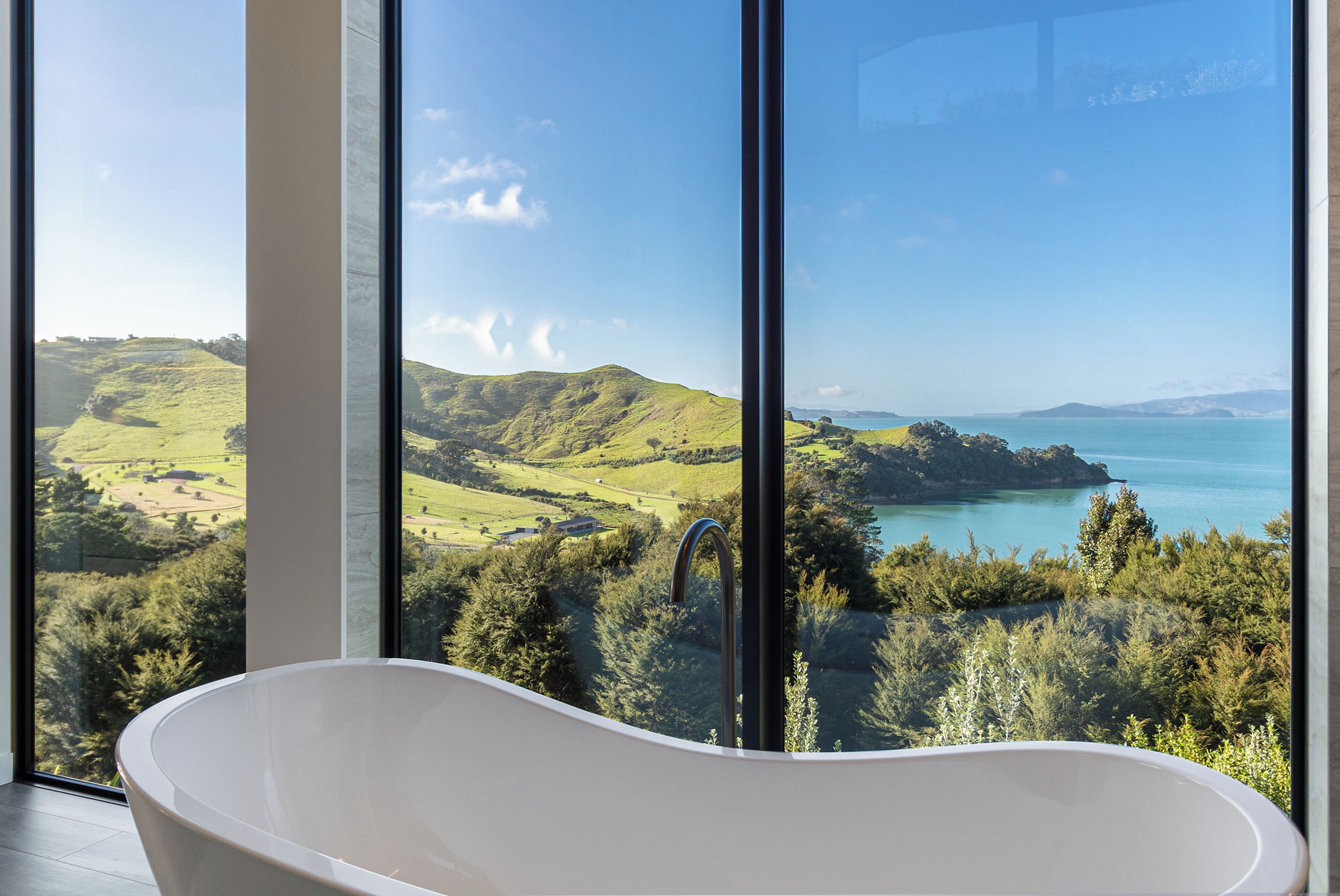 Ein modernes Badezimmer mit freistehender Badewanne und großen Fenstern, die einen schönen Ausblick auf sanfte Hügel und ein Gewässer bieten.