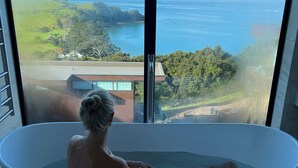 A person in a large bathtub with a view of a coastal landscape through a large window. - Omana (Waiheke Island)