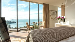 A bedroom with a large bed, a view of the sea, and a dining area with a table and chairs. - Omana (Waiheke Island)