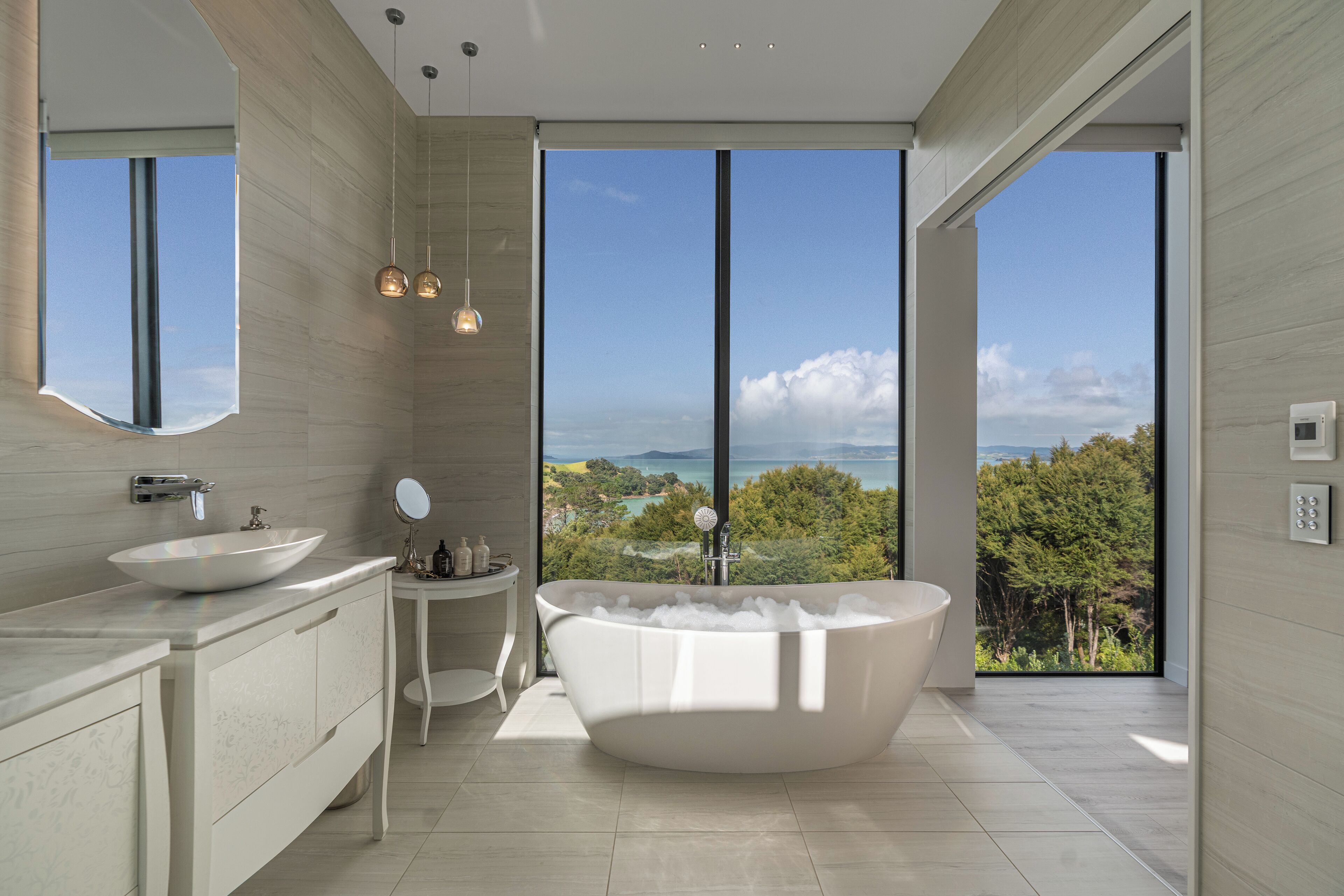 A modern bathroom with a freestanding bathtub, a large mirror, and a view of the sea.