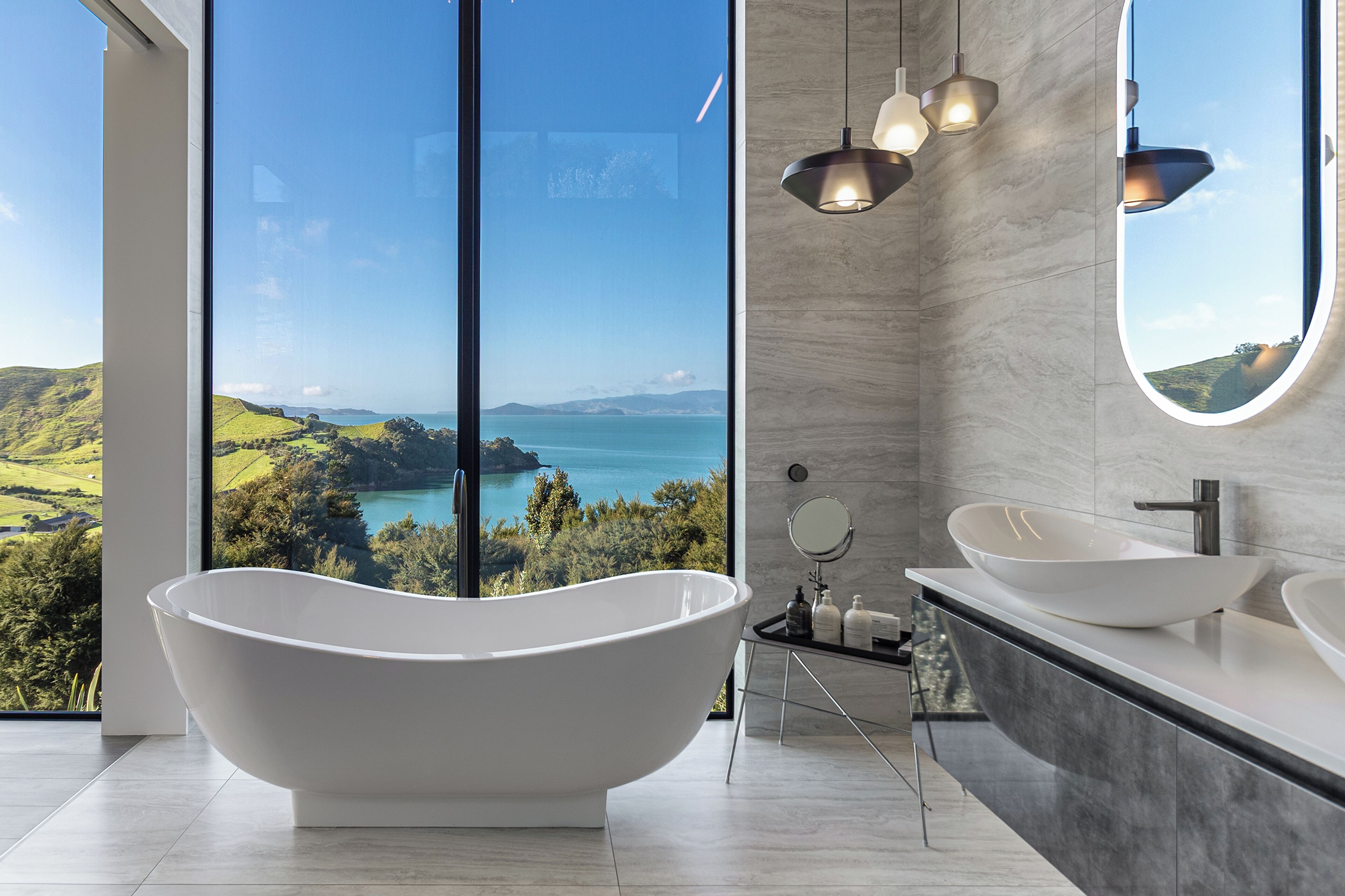 A modern bathroom with a freestanding bathtub, two sinks, a large mirror, and a view of a coastal landscape.