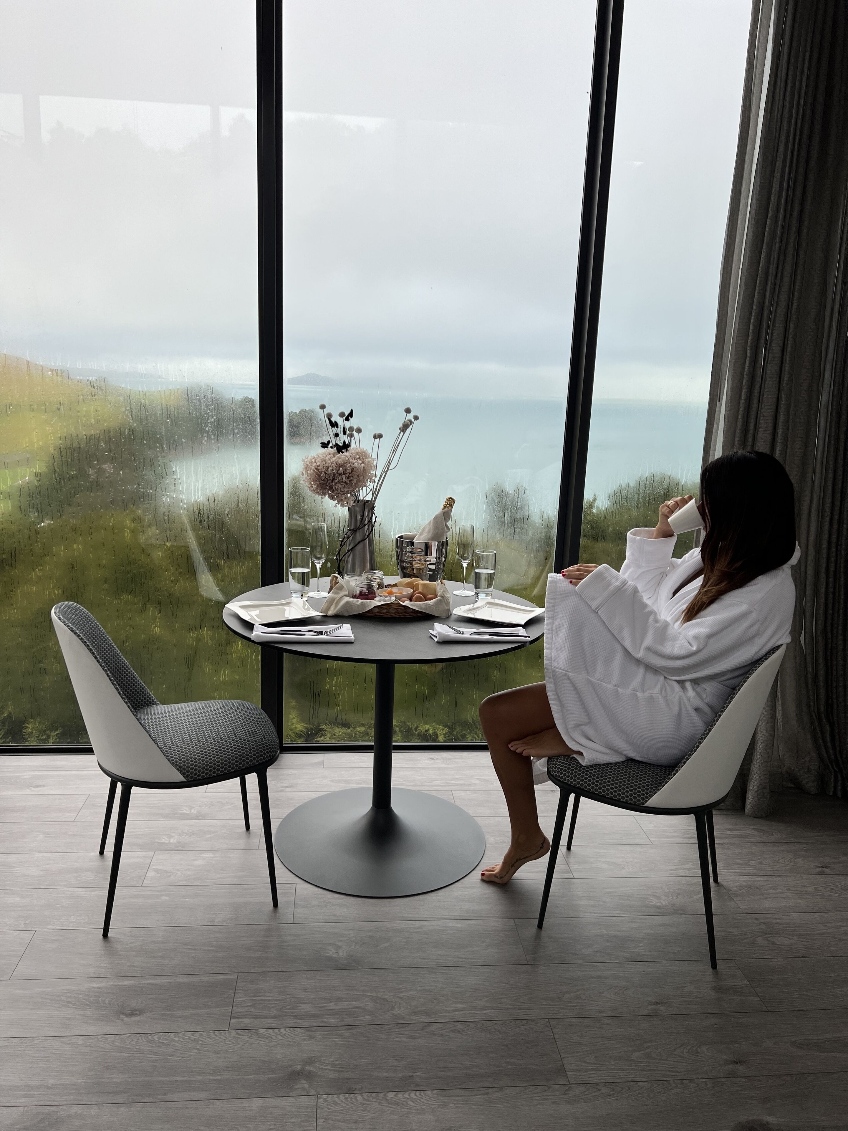 Eine Frau sitzt an einem runden Tisch mit Blick auf einen See und viel Grün. Der Raum ist modern eingerichtet und verfügt über ein großes Fenster, einen Stuhl und einen Tisch mit einer Flasche, Gläsern sowie einem Brotkorb.