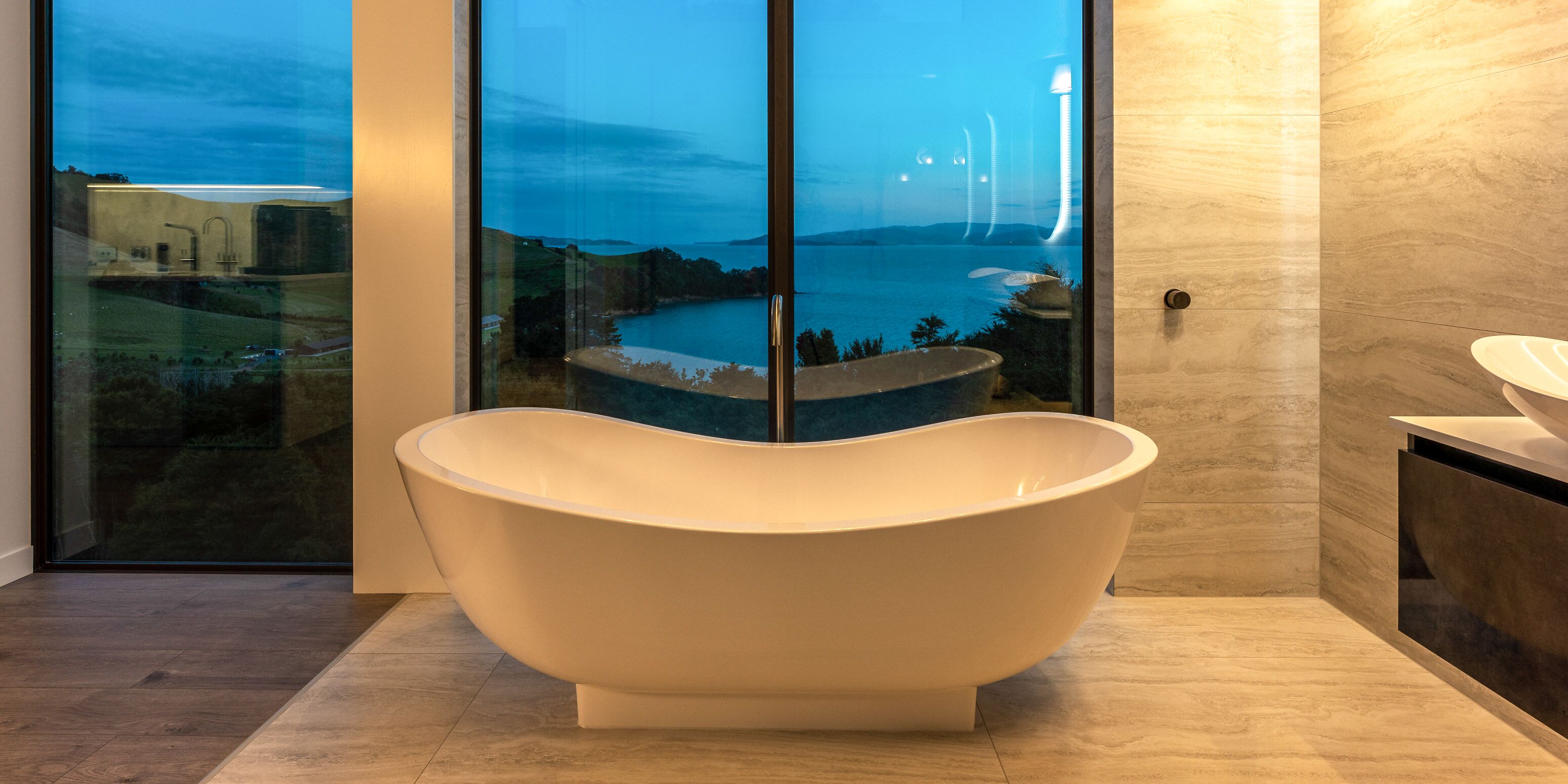 Een moderne badkamer met een vrijstaand bad, grote glazen deuren en uitzicht op een waterlichaam.