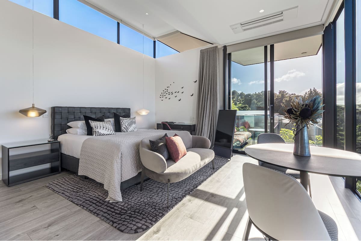 Ein modernes Schlafzimmer mit einem großen Bett, einem Sofa, einem Esstisch und Blick ins Freie.