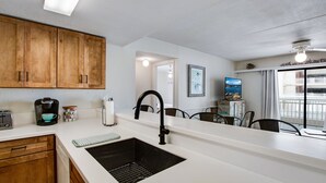 Una cocina con armarios de madera, un lavamanos negro, una cafetera y un comedor con sillas y una mesa.