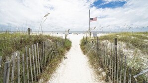 Un sendero arenoso que lleva a una playa con una bandera estadounidense, rodeado de cercas de madera y césped.