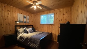 Une chambre avec des murs en bois, un lit avec une tête de lit, un ventilateur de plafond et une fenêtre avec des stores.