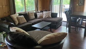 Un salon chaleureux avec un canapé d’angle, une table basse et une table à manger avec des chaises. De grandes fenêtres offrent une vue sur l’extérieur.